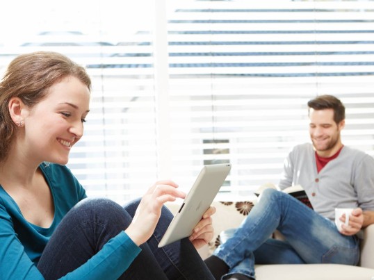 A woman using a tablet and a man reading a book while sitting on a couch.